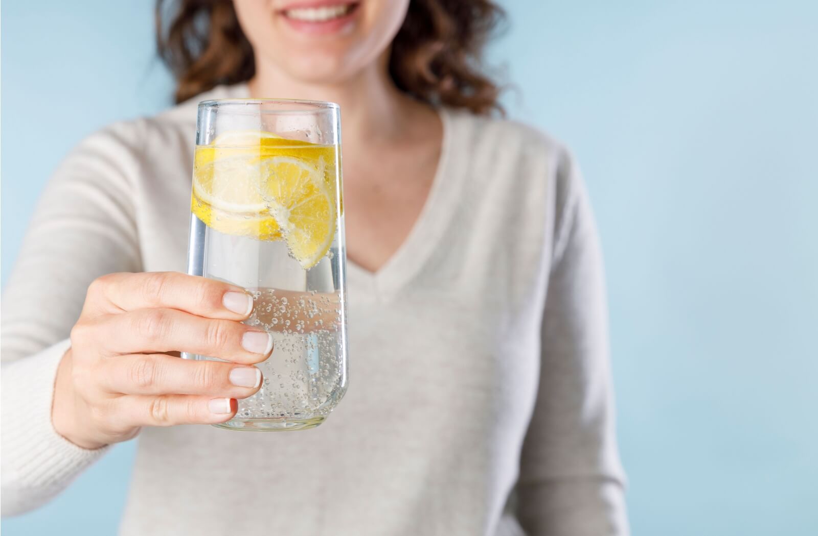 A person standing against a blue background, smiling and holding a glass of sparkling water with lemon slices.