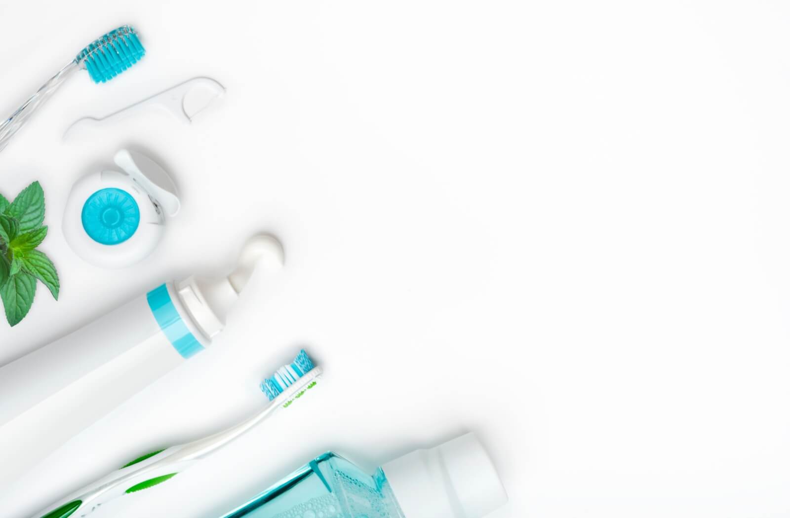 Various dental hygiene tools lay on a white background in minty colours, alongside a sprig of mint itself.